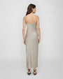 A person shown from the back wearing a long, fitted, light taupe satin‑finish dress with thin spaghetti straps. The dress has a smooth, straight silhouette that falls to the ankles. The individual’s arms rest naturally at the sides, and a pair of strappy, pointed flat shoes is visible.