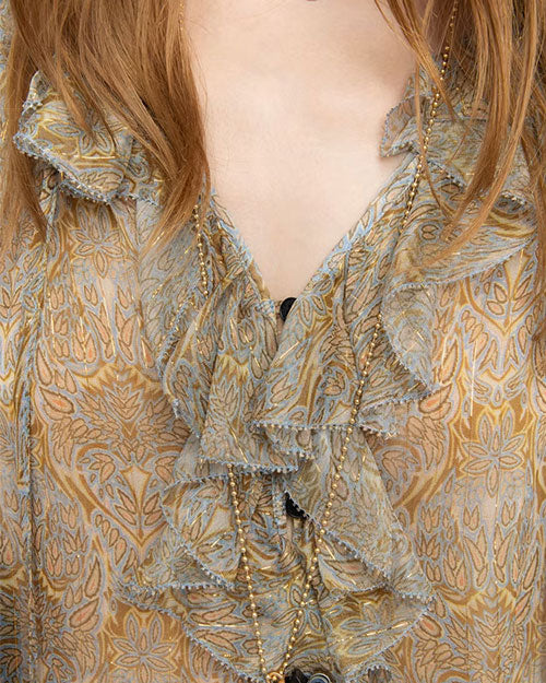Close-up of a person's upper torso wearing a patterned blouse with ruffled neckline and floral and leaf motifs in beige, gold, and blue. A long beaded necklace is draped around the neck, and hair partially covers the shoulders.