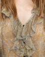 Close-up of a person's upper torso wearing a patterned blouse with ruffled neckline and floral and leaf motifs in beige, gold, and blue. A long beaded necklace is draped around the neck, and hair partially covers the shoulders.