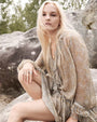 Person with long blonde hair sitting on a large rock outdoors, wearing a light-colored, patterned long-sleeve dress and multiple rings on the left hand. There are trees and rocks in the background.