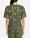 A backside view of a person standing and wearing a matching green outfit with intricate patterns. The outfit consists of short-sleeved top and matching shorts, both adorned with floral and geometric designs in white, blue, and purple colors. The background is plain white.