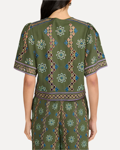 A backside view of a person standing and wearing a matching green outfit with intricate patterns. The outfit consists of short-sleeved top and matching shorts, both adorned with floral and geometric designs in white, blue, and purple colors. The background is plain white.
