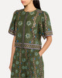 A close-up body shot of a dark-haired model with her hand in her pocket, standing against a white background and wearing a matching green short-sleeved top and bottoms with intricate patterns, adorned with floral and geometric designs. 