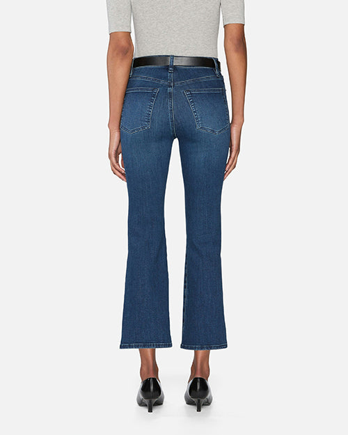 The back view of a model wearing a pair of blue denim jeans with a black belt. The jeans are high-waisted and slightly flared at the bottom, with two back pockets and frayed hems.  They are paired with black pointed-toe shoes. The person is also wearing a light grey top with short sleeves. The background is plain white.