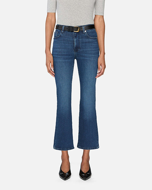 A model is wearing a pair of blue denim jeans with a black belt. The jeans are high-waisted and slightly flared at the bottom, featuring two large front pockets and frayed hems. The denim has a slightly worn and faded look, particularly around the knees and thighs. They are paired with black pointed-toe shoes. The person is also wearing a light grey top with short sleeves. The background is plain white.
