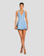 A full body image of a brunette model wearing a sleeveless, short light blue denim dress with a deep V-neckline and vertical seam work. She is accessorized with gold high-heeled sandals, gold bracelets, a gold handbag, and a gold choker necklace. The background is plain gray.