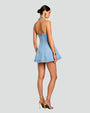 A full body backside image of a brunette model in a bun wearing a halter top, open back, short light blue denim dress with vertical seam work. She is accessorized with gold high-heeled sandals, gold hoop earrings and a gold bracelet. background is plain grays.