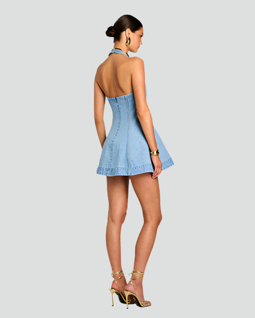A full body backside image of a brunette model in a bun wearing a halter top, open back, short light blue denim dress with vertical seam work. She is accessorized with gold high-heeled sandals, gold hoop earrings and a gold bracelet. background is plain grays.
