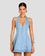 A knee-up image of a brunette model wearing a sleeveless, short light blue denim dress with a deep V-neckline and vertical seam work. She is accessorized with gold hoop earrings, gold bracelets, and a gold choker necklace. The background is plain gray.