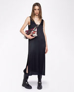 A brunette model wearing a long, sleeveless black dress with a deep V-neckline and lace detailing around the chest and arm openings. She is holding a colorful handbag and chunky black boots, while standing against a white background.