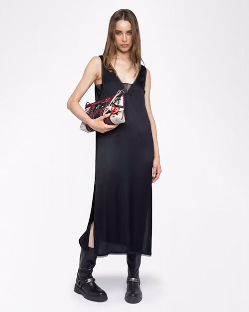 A brunette model wearing a long, sleeveless black dress with a deep V-neckline and lace detailing around the chest and arm openings. She is holding a colorful handbag and chunky black boots, while standing against a white background.