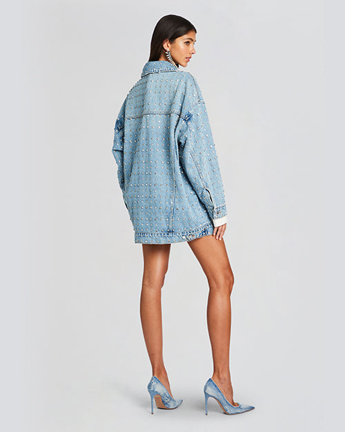 A back view of a model wearing an oversized light blue denim jacket with rhinestones all over. The jacket features a classic collar. The sleeves are long and cuffed at the wrists. Model is styled with denim high heels.