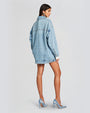 A back view of a model wearing an oversized light blue denim jacket with rhinestones all over. The jacket features a classic collar. The sleeves are long and cuffed at the wrists. Model is styled with denim high heels.