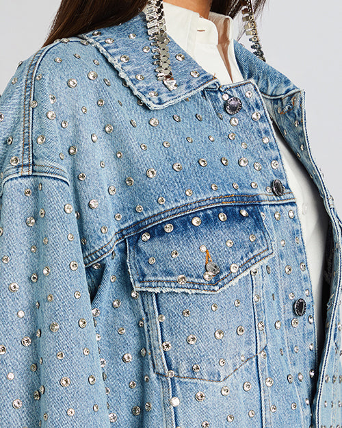 A close up of a model wearing an oversized light blue denim jacket with rhinestones all over. The jacket features a classic collar, silver hardware, buttoned chest pockets, and side pockets. 