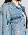 A close up of a model wearing an oversized light blue denim jacket with rhinestones all over. The jacket features a classic collar, silver hardware, buttoned chest pockets, and side pockets. 