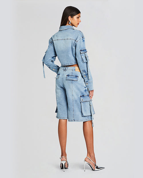 Back view of a model wearing a cropped denim long sleeved top. The top features a classic collar, a front button closure and cargo pockets with draping adjustable straps. The model is styled with matching denim shorts and silver heels.