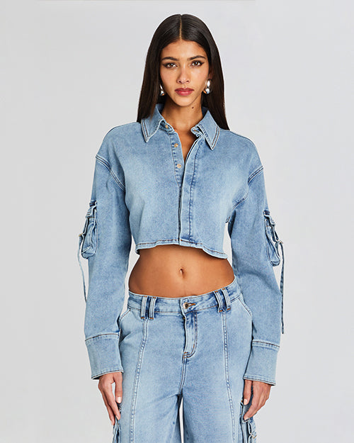 A model wearing a cropped denim long sleeved top. The top features a classic collar, a front button closure. The sleeves have cargo pockets with draping adjuistable straps. The model is styled with matching denim shorts.