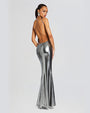 A backside full body view of a brunette model wearing a floor-length, metallic silver dress with a backless design and thin straps. The model is wearing silver earrings and is standing in front of a white background.
