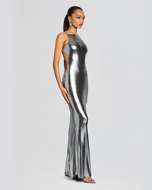 An angled full body view of a brunette model wearing a floor-length, metallic silver dress with a backless design and thin straps. The model is wearing silver earrings, open-toed heels and is standing in front of a white background.