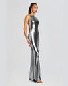 A side view of a model wearing a floor-length, metallic silver dress with a high straight neckline and a form-fitting silhouette. The back of the dress appears to have a low back design. The dress has a shimmering effect. The model is styled with silver accessories.