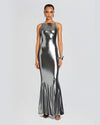 A full view of a model wearing a floor-length, metallic silver dress with a high straight neckline and a form-fitting silhouette. The dress has a shimmering effect. The model is styled with silver accessories.