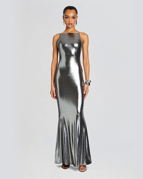 A full body view of a brunette model wearing a floor-length, metallic silver dress with thin straps and an accordion skirt design. The model is wearing silver earrings, open-toed heels and is standing in front of a white background.