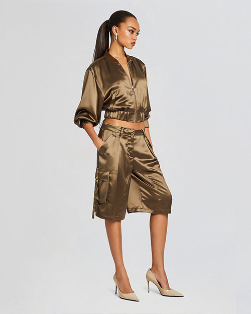 A full view of a model wearing a cropped olive green satin bomber jacket with a front silver zipper and gathered elastic hem and cuffs. The person is also wearing matching satin shorts.