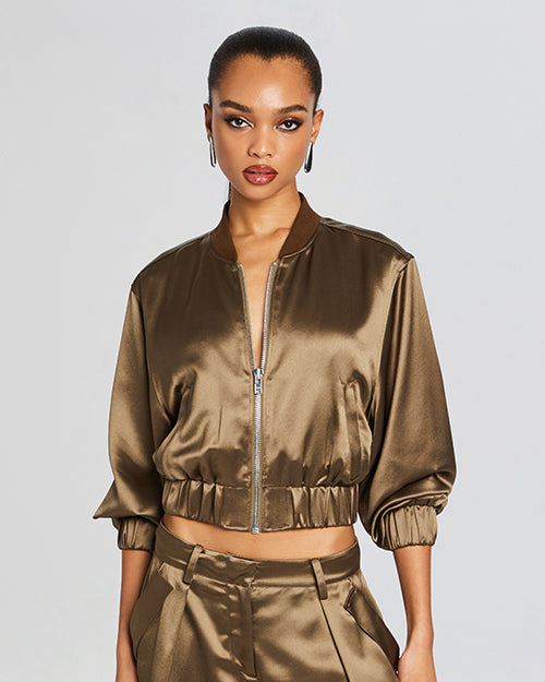 A model wearing a cropped olive green satin bomber jacket with a front silver zipper and gathered elastic hem and cuffs. The person is also wearing matching satin shorts.