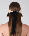 Back view of a person with long, straight brown hair wearing a beige straw hat adorned with a wide black ribbon tied into a bow, with the ribbon ends hanging down the back. The background is plain and light-colored.