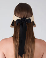 Back view of a person with long, straight brown hair wearing a beige straw hat adorned with a wide black ribbon tied into a bow, with the ribbon ends hanging down the back. The background is plain and light-colored.