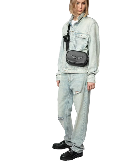 A person wearing a light blue denim jacket and matching ripped jeans, black shoes, and carrying a black crossbody bag with an emblem. 