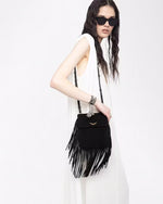 A knee-up view of a dark-haired model wearing a sleeveless white dress and carrying a black over the shoulder handbag with tassels at the bottom and sides, and gold wings at the top center. The model is also wearing black sunglasses and standing against a plain white background.