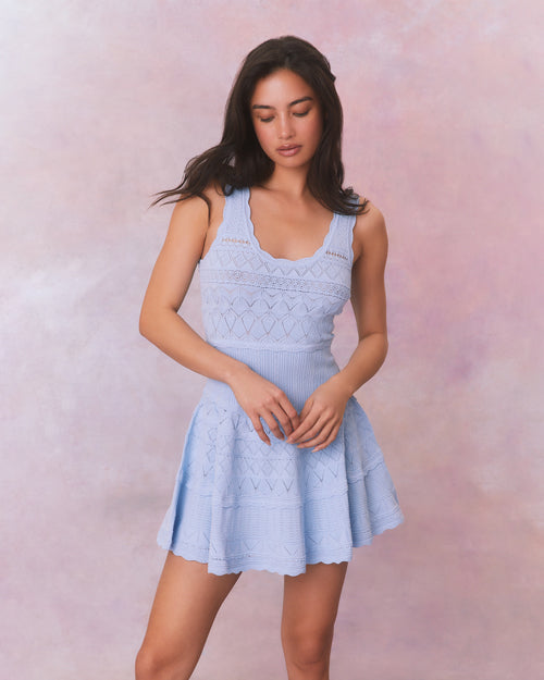 A light blue dress with a fitted bodice and flared skirt, featuring delicate ruffle details, a zigzag lace pattern, and vertical ribbing.