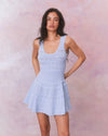 A model in sleeveless, light blue dress with a fitted bodice and a flared skirt. The dress features ruffle details along the straps and neckline, with mesh zigzag pattern and ribbing throughout. 