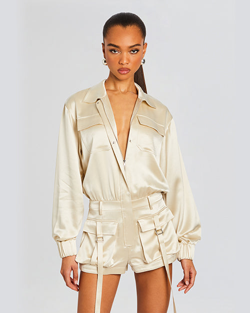 A knee-up view of a model wearing a beige satin shirt with a V-neck collar and matching shorts, with utility style pockets and flaps on both front sides and long strings. The background is white.