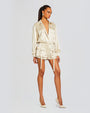 Model wears a beige satin romper with a collared jacket top and shorts. The romper has utility detailing, low V neck, pockets with flaps on both sides. The outfit features a belted waist, pockets below the waist with long straps.