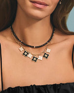 Close-up of an individual’s neck and upper chest area, wearing a black  top. Person wearing two necklaces. Short necklace had black bead stones, while longer is a gold chain with seven Moroccan flowers in gold and black color.