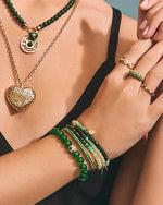 A close-up image of a person’s upper body showing their arms and hands adorned with various pieces of jewelry. On the neck, there is a gold chain necklace with a heart-shaped pendant, and another necklace with round green beads and a circular pendant. The left wrist has several bracelets including green beaded ones, gold bangles, and one with small charms.