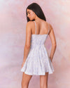 A back view of a model in a short white dress with a subtle blue floral pattern. The dress features thin straps, a fitted bodice with a sweetheart neckline, and flares out into an A-line skirt.
