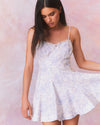 A close-up of a model in a short white dress with a subtle blue floral pattern. The dress features thin straps, a fitted bodice with a sweetheart neckline, and flares out into an A-line skirt.