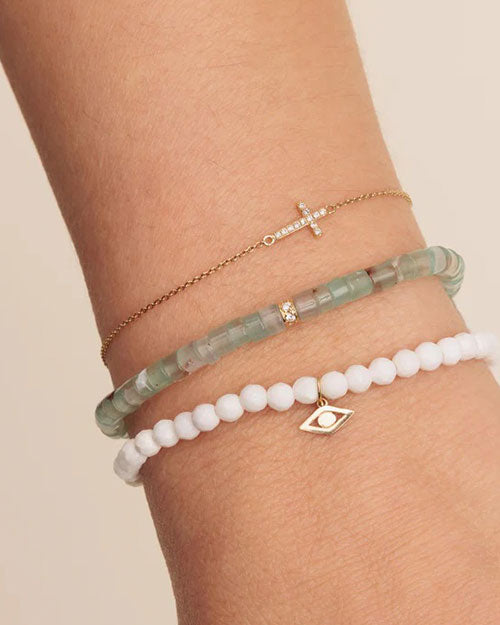 A close-up image of a person’s wrist adorned with three bracelets. The top bracelet is a delicate chain with a small cross charm. The middle bracelet features round greenish stones, with a single gold-colored bead. The bottom bracelet is made of white beads and has a gold  charm in the shape of an eye.