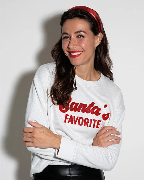 A person is wearing a white sweatshirt with the phrase ‘Santa’s Favorite’ printed in red on the front. The individual is also wearing black leather pants and a red headband. 