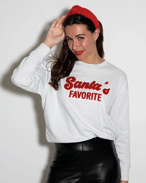 A person is wearing a white sweatshirt with the phrase ‘Santa’s Favorite’ printed in red on the front. The individual is also wearing black leather pants and a red headband. 