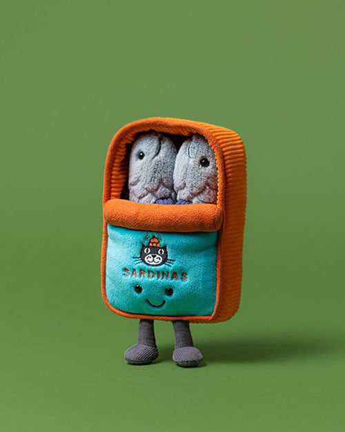 Plush toy resembling a can of sardines with an orange lid and base, two gray sardine plushies inside, turquoise front featuring a cat illustration and the word 'SARDINAS', plus small black eyes and a smiling mouth. Set against a solid green background.