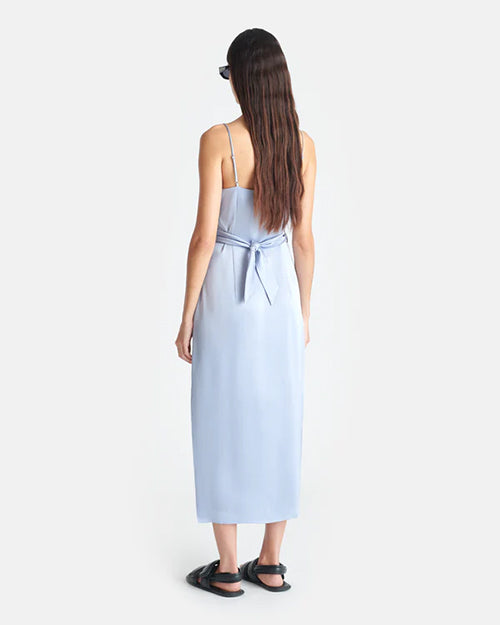 The backside view of a model wearing a light blue shin length dress with thin straps. The dress has light blue fabric tied around the waist. The person is also wearing black sandals and sunglasses to match.