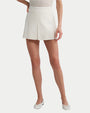 A person is wearing white short shorts with a front pleat. The person is also wearing a white top tucked into the skirt and white flat shoes with black stripes. The background is plain white.