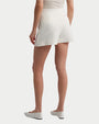 The back view of a person wearing white short shorts with a front pleat. The person is also wearing a white top tucked into the shorts and white flat shoes with black stripes. The background is plain white.