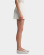 The side view of a person wearing white shorts with a front pleat. The person is also wearing a white top tucked into the shorts and white flat shoes with black stripes. The background is plain white.