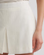 A close-up view of a person wearing white short shorts with a front pleat. The person is also wearing a white top tucked into the shorts and white flat shoes with black stripes. The background is plain white.