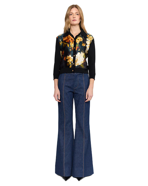 Person standing and facing forward, wearing a black long-sleeve shirt with a vibrant floral print in yellow, white, and orange. Paired with high-waisted, wide-leg denim jeans featuring prominent vertical seams.